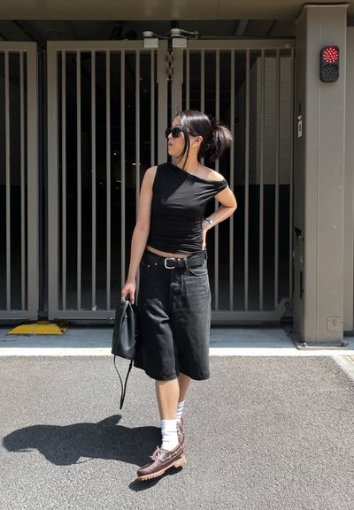 Top negro de un solo hombro y culottes de mezclilla oscuros, combinados con zapatos marrones de cordones y calcetines blancos. Sostiene un bolso negro. Fondo estructurado.