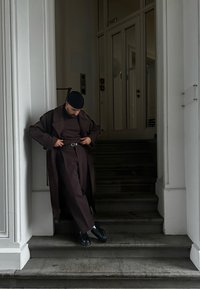 Καφέ παλτό και αντίστοιχες παντελόνες, μαύρη ζώνη, σκούρα παπούτσια. Στεκόμενος σε σκαλοπάτια κοντά σε έναν λευκό τοίχο, ρυθμίζει το παλτό.