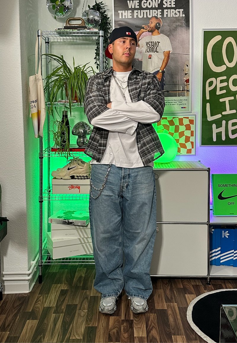 Tenue décontractée comprenant une chemise à carreaux sur un T-shirt blanc, un jean baggy bleu clair et des baskets grises. Les accessoires incluent une chaîne en argent.
