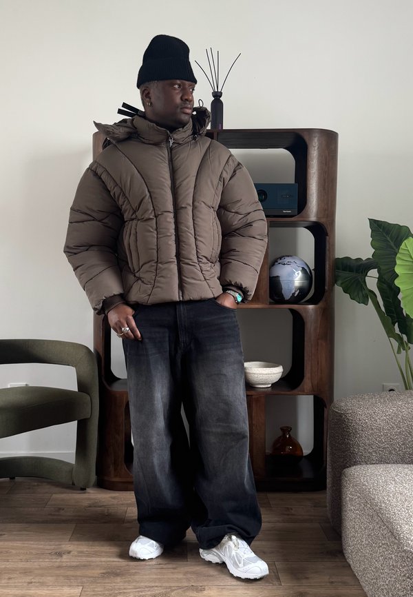 Mann in brauner Steppjacke, schwarzer Mütze und weitem schwarzen Jeans steht drinnen neben einem hölzernen Regal mit Globus und Pflanzen.