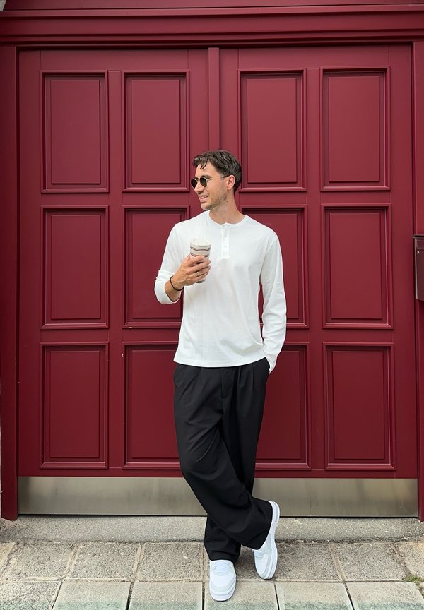 Homme portant des lunettes de soleil, une chemise blanche à manches longues, un pantalon noir et des baskets blanches, tenant une boisson debout devant une grande porte rouge à panneaux.