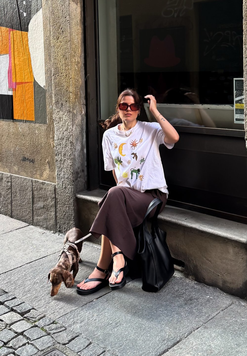 Uma mulher está sentada numa escada de pedra, vestindo uma t-shirt gráfica branca e uma saia castanha, com sandálias pretas. Um pequeno cão preso à trela está ao seu lado.