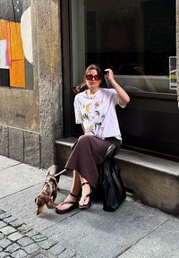 Uma mulher está sentada numa escada de pedra, vestindo uma t-shirt gráfica branca e uma saia castanha, com sandálias pretas. Um pequeno cão preso à trela está ao seu lado.