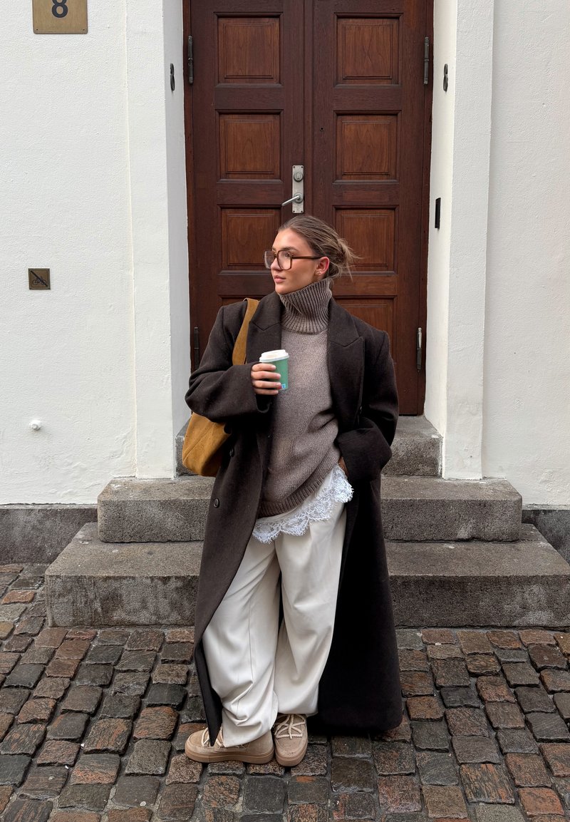 Femme portant un manteau sombre surdimensionné, pull à col roulé beige, pantalon blanc, lunettes, portant un sac marron, tenant une tasse de café verte, debout sur des marches pavées.