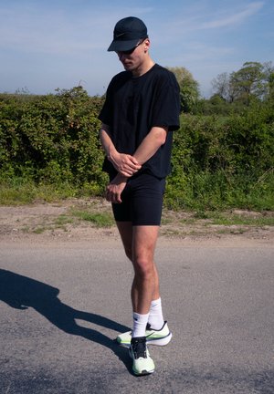 Joven vestido con ropa deportiva negra, gorra y gafas de sol, de pie en la carretera junto a la vegetación en un día soleado.