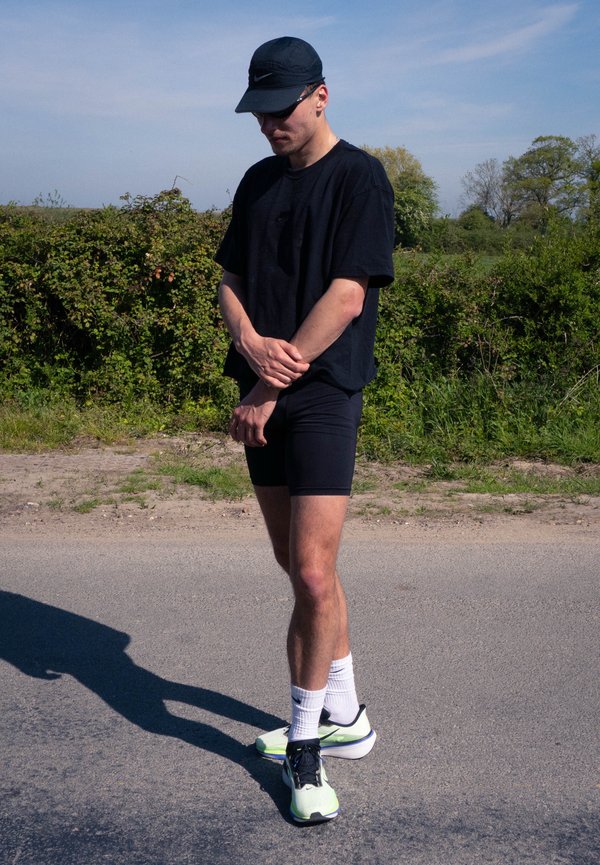 Nuori mies mustassa urheiluasussa, lippalakki ja aurinkolasit päällä, seisoo tiellä vihreän äärellä aurinkoisena päivänä.