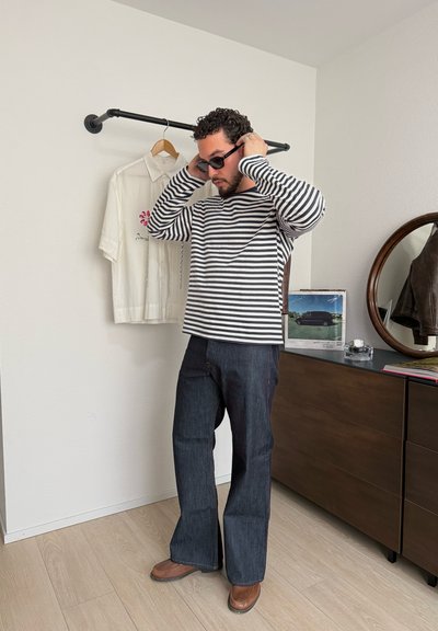 Hombre con gafas de sol negras, camiseta de manga larga a rayas en blanco y negro, vaqueros azules y zapatos marrones en una habitación con camisa blanca colgada y cómoda de madera.