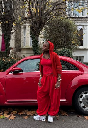 Cardigan de punto rojo con capucha, camiseta sin mangas roja y pantalones rojos holgados. Zapatillas blancas con diseño texturizado. Junto a un coche rojo en un entorno al aire libre.