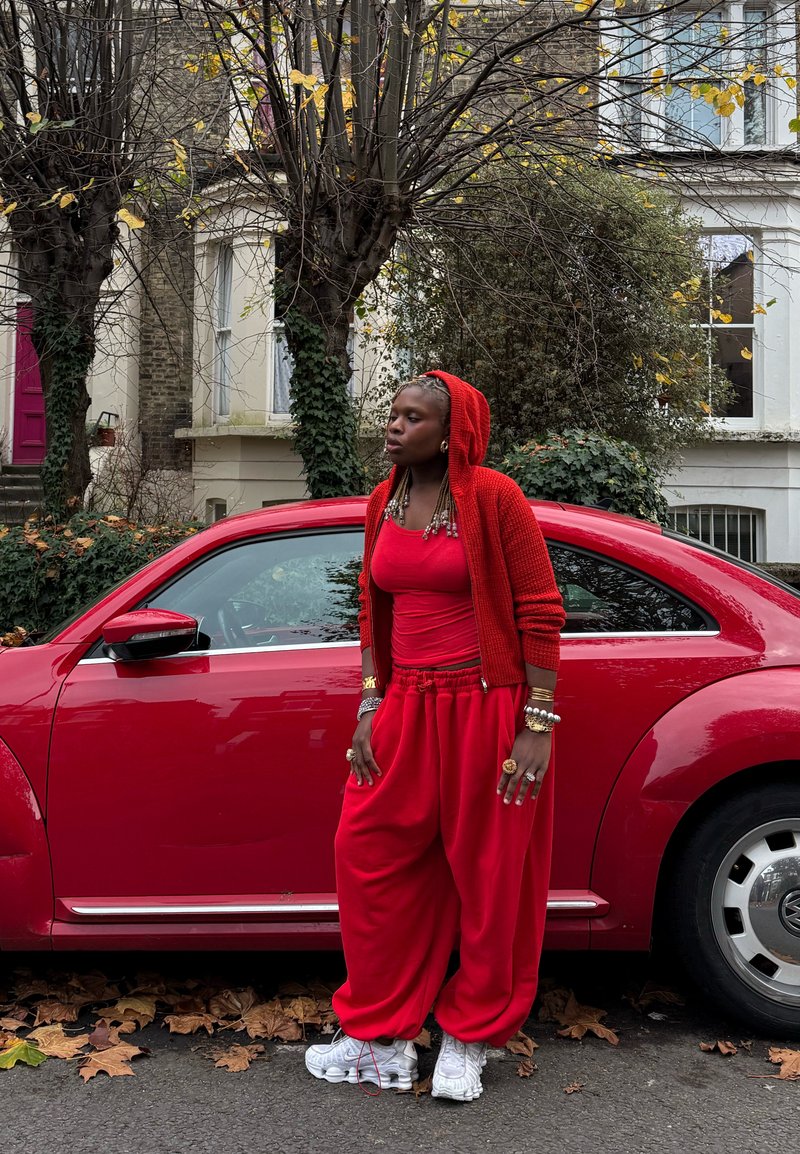 Cardigan lavorato a maglia rosso con cappuccio, canottiera rossa e pantaloni rossi larghi. Scarpe da ginnastica bianche con design testurizzato. Accanto a un'auto rossa in un ambiente esterno.