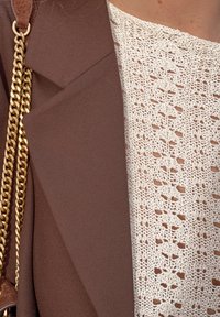 Blazer en tissu marron avec un accent en chaîne, associé à un haut blanc en crochet présentant un motif de feuilles et une texture ajourée.