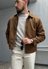 Καφέ σουέτ τζάκετ με φερμουάρ μπροστά, κρέμα φούτερ από μέσα και ανοιχτόχρωμο παντελόνι. Τα αξεσουάρ περιλαμβάνουν γυαλιά ηλίου και ρολόι.