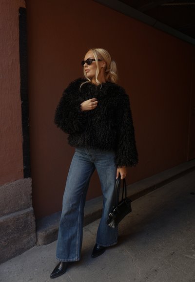 Chaqueta de piel sintética negra y esponjosa, vaqueros azules de pierna ancha, zapatos negros puntiagudos y un pequeño bolso negro. Color sólido, textura suave.