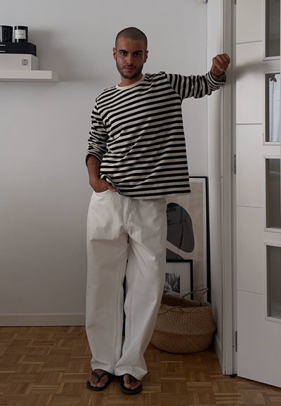Camisa de manga larga a rayas en blanco y negro, pantalones blancos holgados y chanclas negras; de pie junto a una pared con arte y una cesta de almacenamiento.