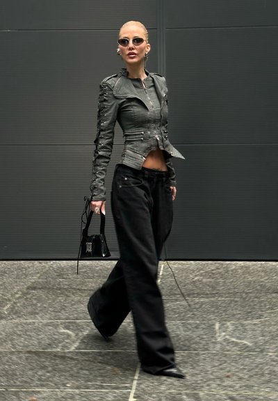 Mujer caminando con pantalones negros anchos, chaqueta de cuero negra ajustada, gafas de sol, sosteniendo un bolso negro pequeño frente a una pared oscura.