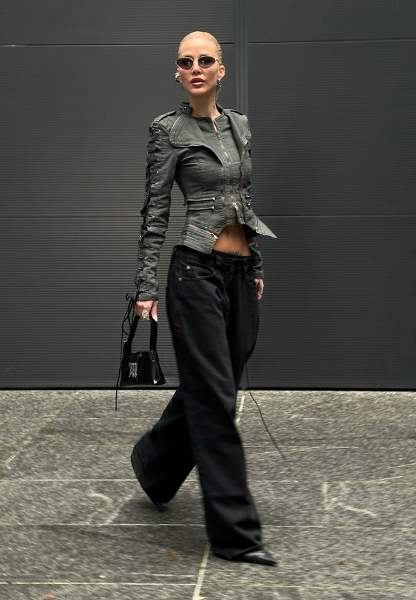 Nainen kävelee mustuissa leveälahkeisissa housuissa, istuvassa mustassa nahkatakissa, aurinkolaseissa ja pitää kädessään pientä mustaa käsilaukkua tummaa seinää vasten.