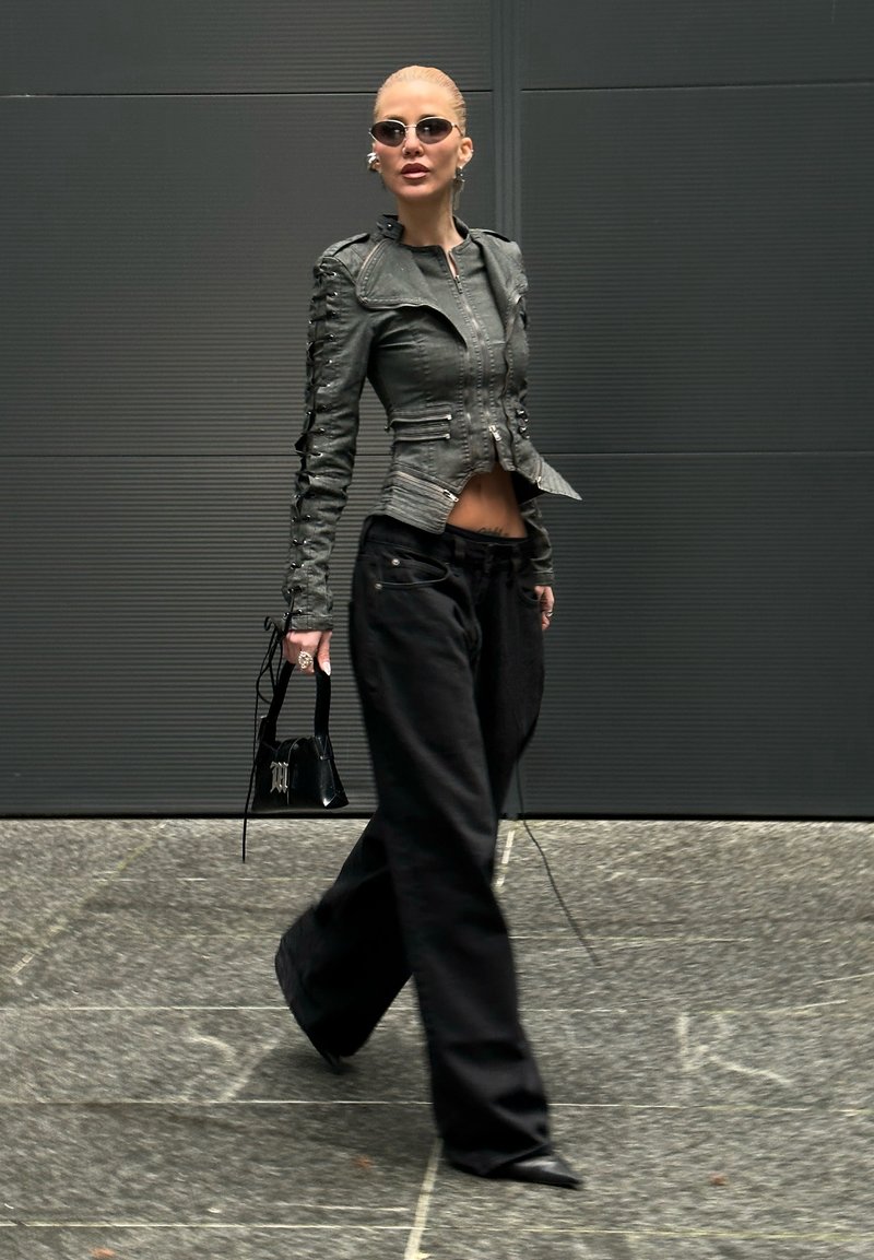 Femme marchant en pantalon large noir, veste en cuir noire ajustée, lunettes de soleil, tenant un petit sac à main noir devant un mur sombre.