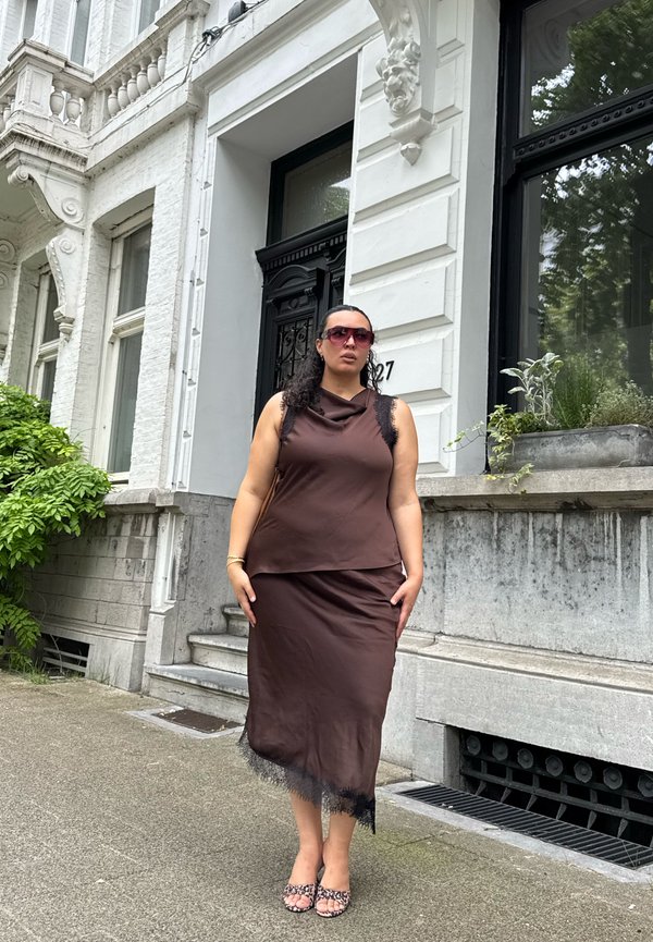 Femme portant un haut marron sans manches et une jupe avec une bordure en dentelle noire, de grandes lunettes de soleil, debout sur le trottoir devant un bâtiment blanc orné.