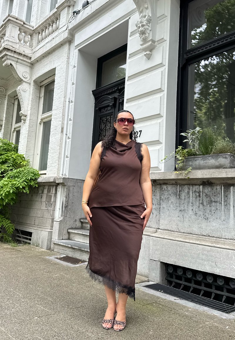 Femme portant un haut marron sans manches et une jupe avec une bordure en dentelle noire, de grandes lunettes de soleil, debout sur le trottoir devant un bâtiment blanc orné.