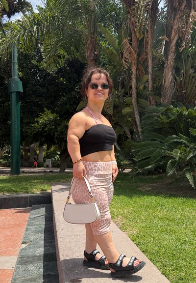 Mujer con gafas de sol, top corto negro, falda estampada y sandalias, de pie al aire libre sosteniendo un bolso blanco con palmeras de fondo.