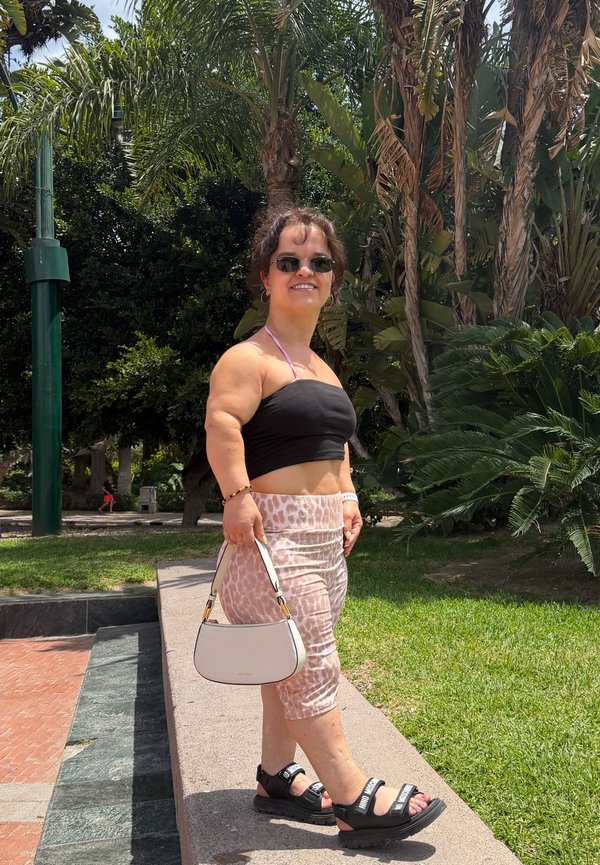 Donna che indossa occhiali da sole, un top corto nero, gonna a motivo e sandali, in piedi all'aperto con una borsa bianca con palme sullo sfondo.