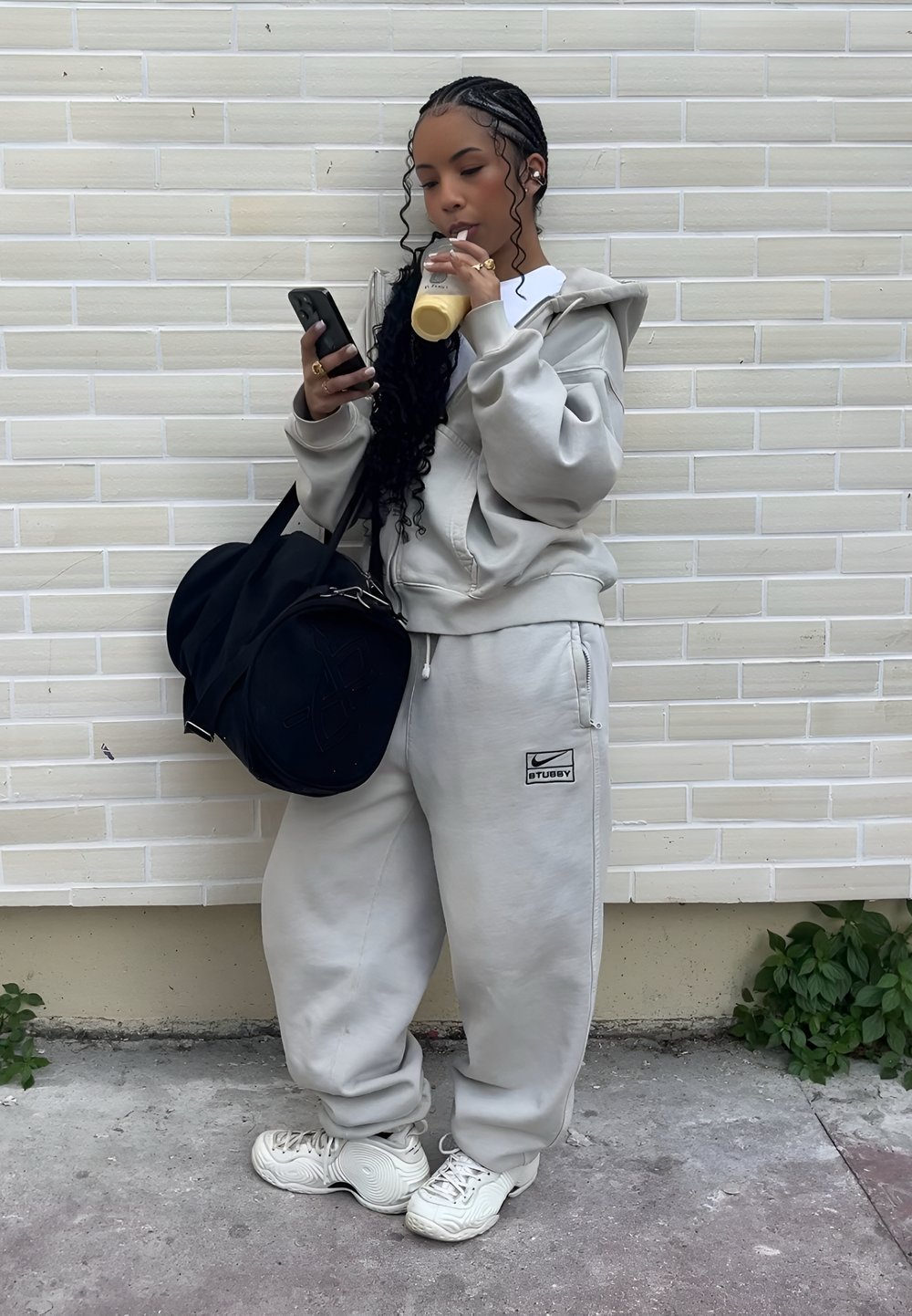 Jeune femme en survêtement gris et baskets blanches boit un smoothie, regarde son téléphone et porte un grand sac de sport noir contre un mur en briques pâles.