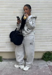 Jeune femme en survêtement gris et baskets blanches boit un smoothie, regarde son téléphone et porte un grand sac de sport noir contre un mur en briques pâles.
