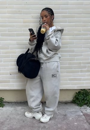 Mujer joven con chándal gris y zapatillas blancas bebe un batido, revisa su teléfono y lleva una gran bolsa de viaje negra frente a una pared de ladrillos pálidos.