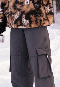 Cargo-broeken van donkergrijze stof, met zijzakken, een gladde textuur en een metalen ringaccent. Gecombineerd met een bruin fleece top met patroon.