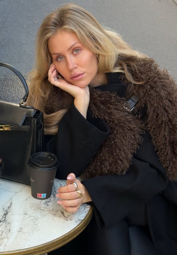 Mujer rubia con ojos verdes apoya la cabeza en la mano en una mesa de mármol con bolso negro y taza de café para llevar al aire libre.
