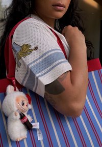 Λευκό διχτυωτό πουκάμισο με μπλε ρίγες και γραφικό λιοντάρι; κόκκινη τσάντα ώμου με κατακόρυφες μπλε και κόκκινες ρίγες; μαλακό παιχνίδι συνδεδεμένο με μπουκάλι Coca-Cola.