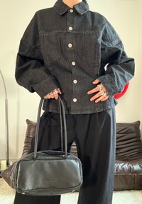 Personne portant une veste en jean sombre surdimensionnée et un pantalon noir, tenant un sac à main en cuir noir avec des poignées à fermeture éclair argentée.