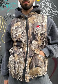 Homme portant un sweat à capuche gris sous une veste camouflage avec le logo Adidas, debout contre un mur couvert de graffiti, une main dans la poche.