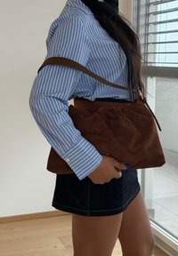 Femme portant une chemise à rayures bleues et une jupe noire tenant un grand sac à main en daim marron avec une bandoulière à l'intérieur.