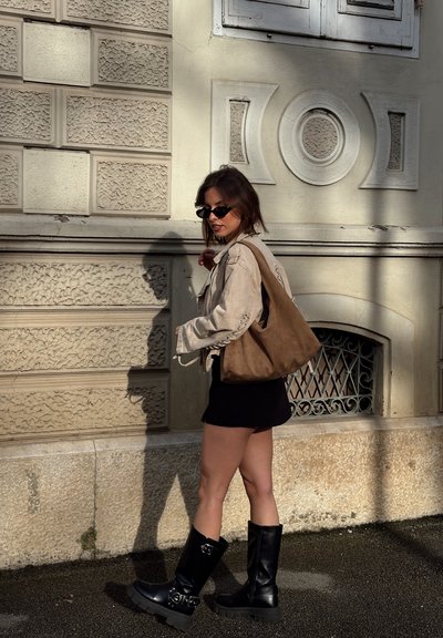 Mujer con chaqueta beige, falda negra, botas altas negras y gafas de sol caminando junto a una pared de piedra texturizada bajo la luz del sol.