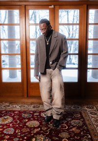 Άνδρας με κοτσίδες και γυαλιά στέκεται σε κόκκινο χαλί με σχέδια, φορώντας γκρι σακάκι, μαύρο πουκάμισο, φαρδιά γκρι τζιν και μαύρα μποτάκια μέσα σε εσωτερικό χώρο.