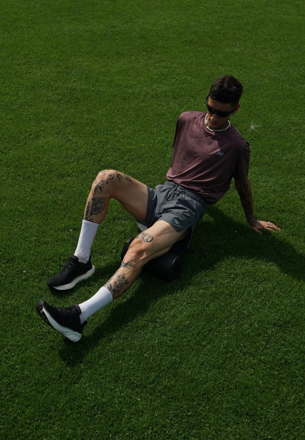 Rodillo de espuma negro sobre hierba, con una persona sentada que lleva una camiseta burdeos, pantalones cortos grises, calcetines blancos y zapatillas negras. Tatuajes visibles en las piernas.