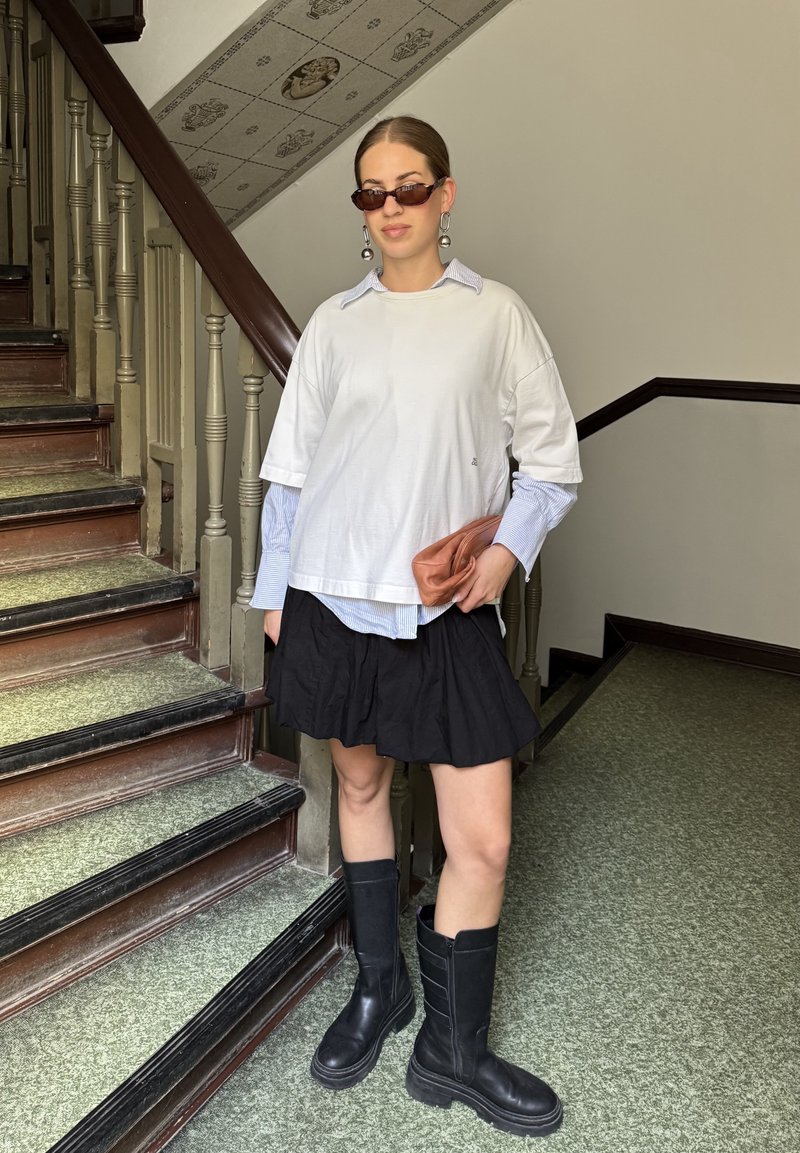 T-shirt blanc surdimensionné superposé à une chemise à col bleu clair, jupe noire et bottes noires jusqu'aux genoux ; tenant une pochette marron.