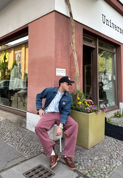 Chaqueta de mezclilla, camisa clara, pantalones rosas, zapatos marrones. Sentado al lado de unos maceteros con flores. Al fondo se puede ver una tienda con grandes ventanas.