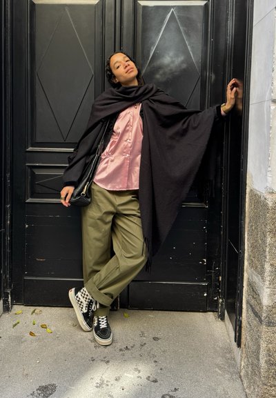 Mujer apoyada contra una puerta negra, vistiendo una camisa rosa de satén, un chal negro, pantalones oliva y zapatillas de cuadros, con una mano levantada.