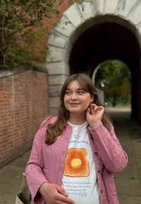 Jonge vrouw in een roze jack die een oorbel aanpast, staande buiten bij een bakstenen muur en stenen boog, glimlachend en naar de zijkant kijkend.