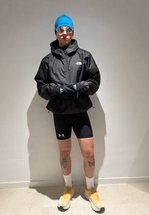 Persona con gorro de baño azul, gafas reflectantes, chaqueta negra de North Face, pantalones cortos negros de Under Armour, guantes negros, calcetines blancos y zapatos amarillos, de pie.