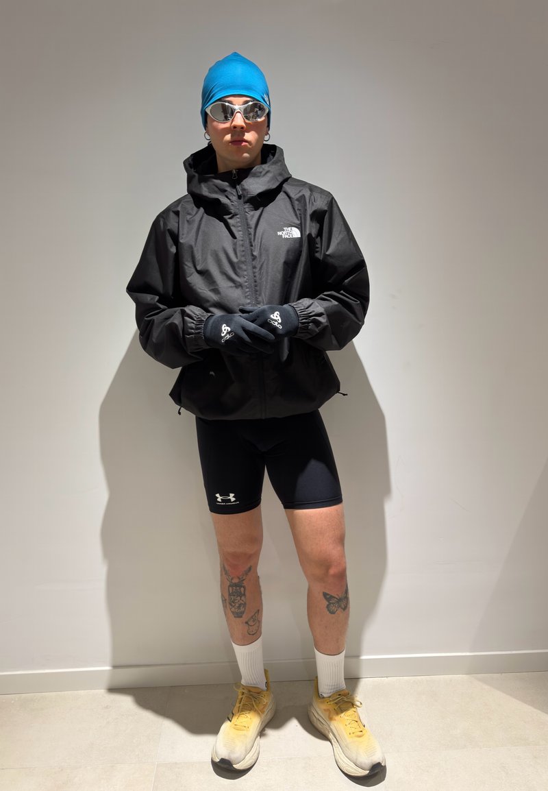 Personne portant un bonnet de bain bleu, des lunettes réfléchissantes, une veste noire North Face, un short noir Under Armour, des gants noirs, des chaussettes blanches et des chaussures jaunes, debout.