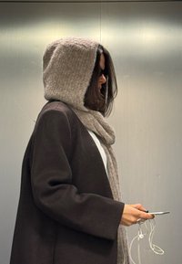 Person i en brun frakk og beige hettehals, som holder en smarttelefon og hvite øretelefoner, og står mot en enkel metallisk bakgrunn.