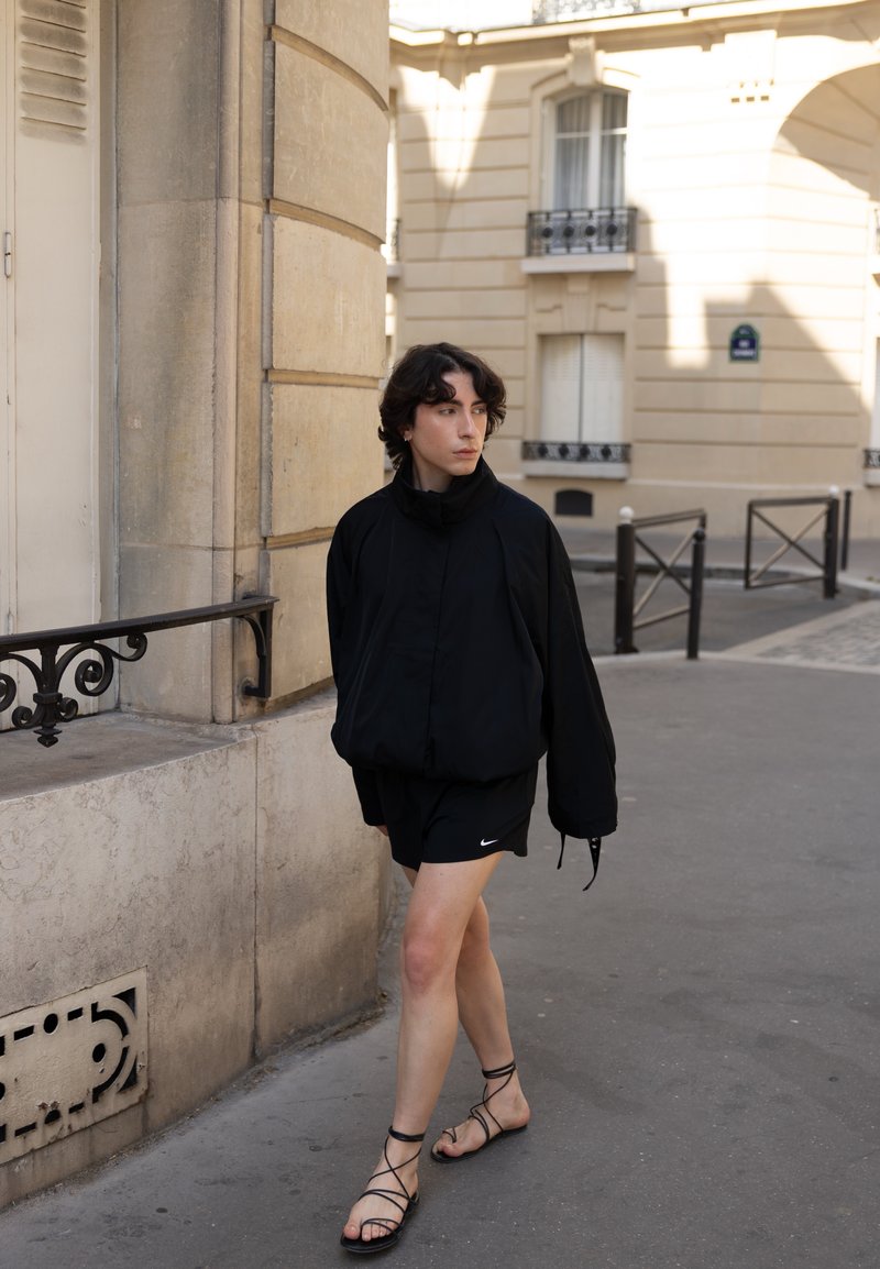 Personne aux cheveux courts et foncés portant une veste noire, un short noir et des sandales noires à brides, marchant sur un trottoir en ville près d'un bâtiment beige.