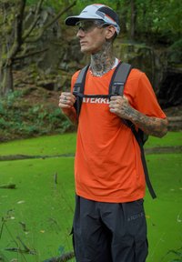 Oranje T-shirt met witte "TERREX" logo, zwarte cargo broek en een zwarte rugzak. Draagt een witte pet en een bril, omringd door groen.