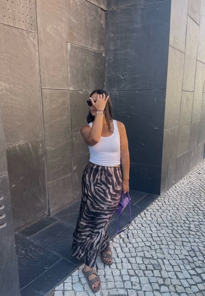 Una persona que lleva una camiseta sin mangas blanca y una falda maxi estampada marrón sostiene un bolso morado, de pie frente a una pared negra texturizada.