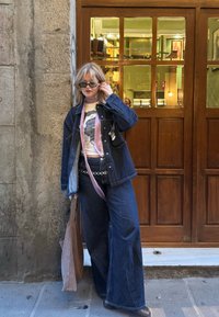 Τζάκετ τζιν με ανοιχτό μπλε λεπτομέρειες πάνω από ένα γραφικό T-shirt. Φαρδιά τζιν παντελόνια σε σκούρο τζιν και ροζ κορδέλα. Καφέ τσάντα tote και μαύρα γυαλία ηλίου.