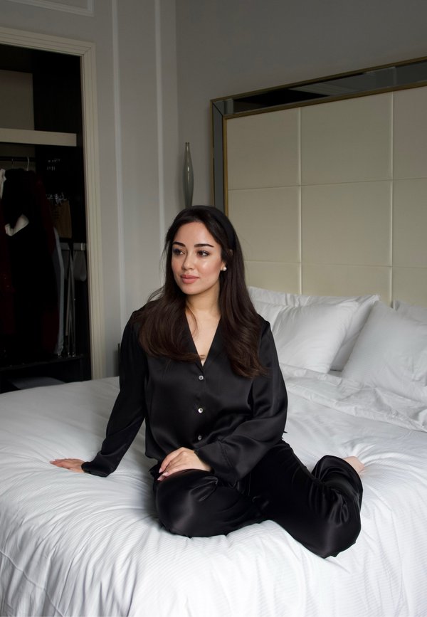 Femme en pyjama de soie noire assise sur un lit blanc dans une chambre avec une tête de lit rembourrée et un placard ouvert en arrière-plan.