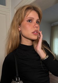 Πορτραίτο ενός ατόμου με μακριά, ίσια ξανθά μαλλιά, φορώντας μια μαύρη εφαρμοστή μπλούζα, ασημένια κρίκους και κρατώντας μια τσάντα με αλυσίδα.