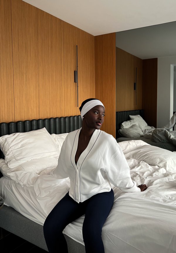 Hvid, let kimono med dyb V-hals og blå kantbånd, båret over mørke leggings, siddende på en seng med krøllet hvidt sengetøj.